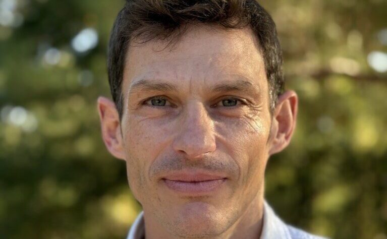 Professional headshot of a man with short, wavy dark brown hair, smiling warmly at the camera. He has fair skin, green eyes, and light stubble, and is wearing a white button-down collared shirt with a thin necklace visible at the neckline. The background is a softly blurred outdoor scene of green foliage, suggesting natural daylight.