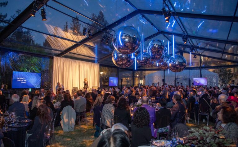 Wide shot of a crowded formal event inside a clear-roofed tent at dusk, titled "Science Under the Stars" as shown on multiple screens around the room. A speaker stands on a small stage in front of a cream curtain, addressing seated guests at round dinner tables. Large mirrored disco-ball sculptures and vertical neon light tubes hang from the ceiling, reflecting the blue evening sky visible through the transparent tent roof, with trees silhouetted in the background.