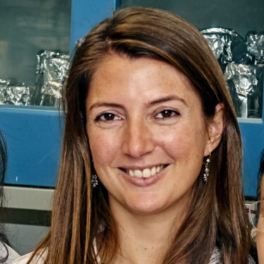 Hheadshot of a woman with long brown hair, smiling warmly at the camera. She has dangling earrings, fair skin, and is wearing a white lab coat over a dark top, with laboratory shelving and foil-covered containers visible in the blurred background.