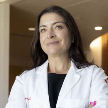 Portrait of a woman with long dark hair standing with her arms crossed, wearing a white lab coat over a black top, gazing thoughtfully off to the side with a confident, composed expression. She's in a modern hallway with warm wood-slat paneling and soft overhead lighting in the background.
