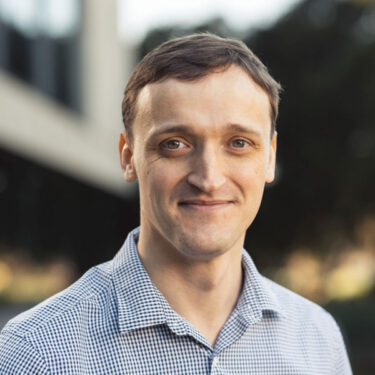 Close-up headshot of a man with short brown hair, smiling softly at the camera. He has fair skin and is wearing a blue-and-white checkered button-down shirt, photographed outdoors with a blurred background of trees and a modern building.