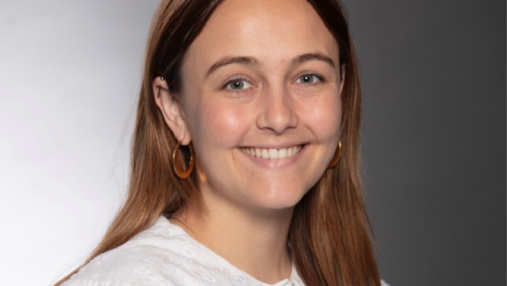 Studio headshot of a young woman with long auburn hair and light eyes, smiling brightly at the camera. She has a small nose stud and gold hoop earrings, and is wearing a white ruffled blouse with an eyelet collar. The background is a softly graduated gray gradient.
