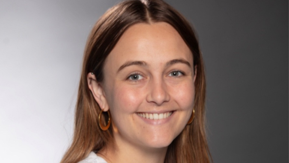Studio headshot of a young woman with long auburn hair and light eyes, smiling brightly at the camera. She has a small nose stud and gold hoop earrings, and is wearing a white ruffled blouse with an eyelet collar. The background is a softly graduated gray gradient.