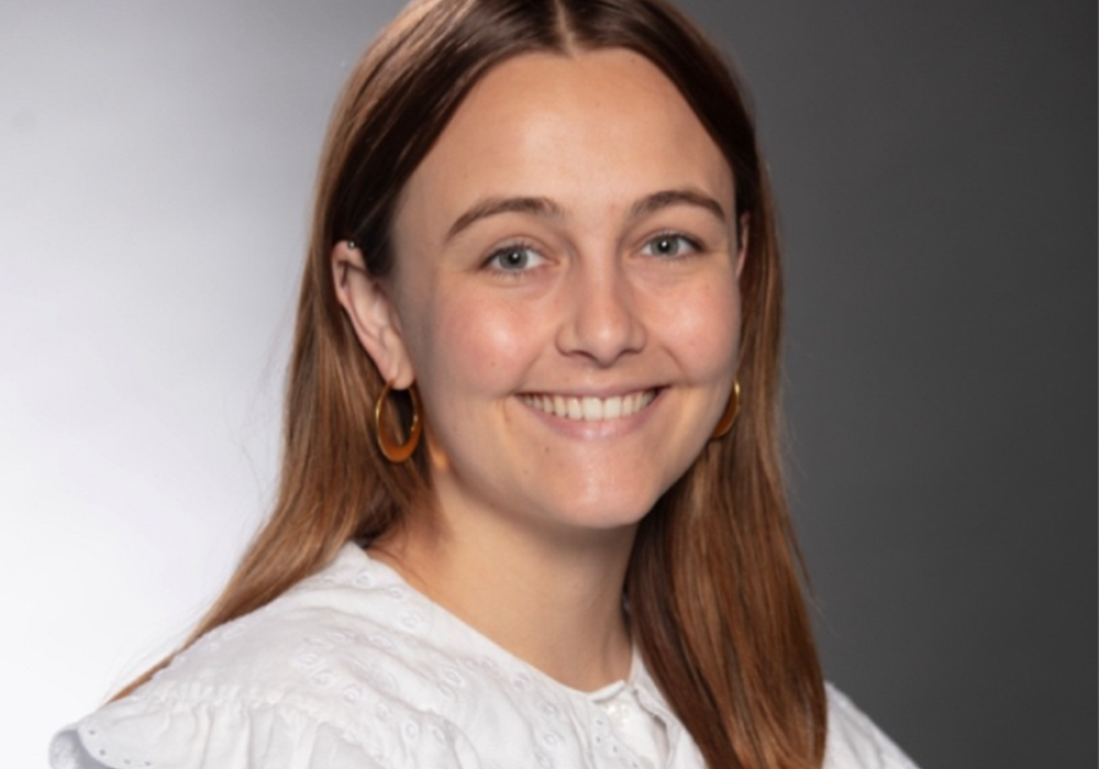 Studio headshot of a young woman with long auburn hair and light eyes, smiling brightly at the camera. She has a small nose stud and gold hoop earrings, and is wearing a white ruffled blouse with an eyelet collar. The background is a softly graduated gray gradient.