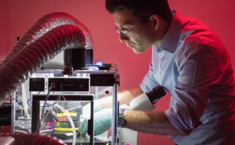 A researcher in a light blue collared shirt and glasses works intently on a piece of lab equipment, lit in red ambient lighting. He wears a blue nitrile glove on his right hand as he adjusts components inside a clear enclosure connected to a large ribbed exhaust tube. The setup includes various dark electronic modules, cables, and a small blue indicator light, suggesting a scientific or materials research experiment in progress.