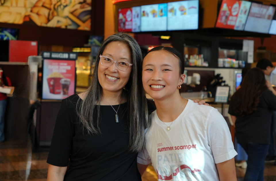 A Pixar production assistant and a former Stanford Children's patient stand together in a movie theater in front of the concession stand, smiling directly at the camera.