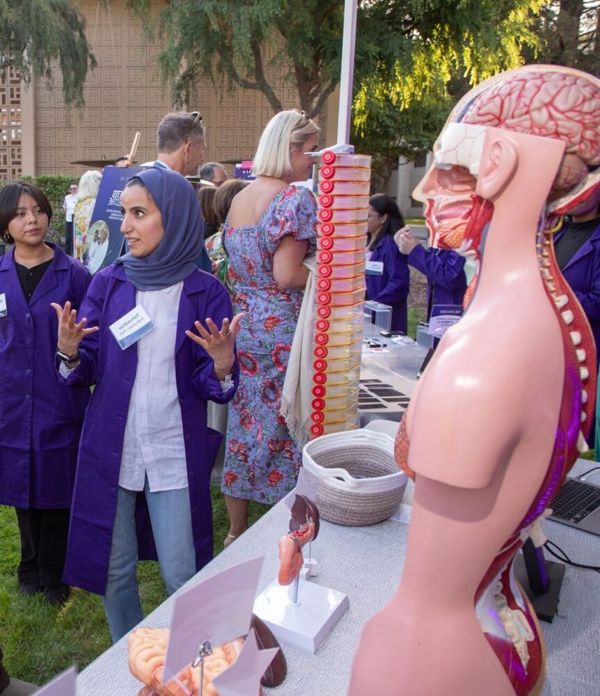 At an outdoor event, a man in a tweed jacket named John Banning talks with two researchers in purple lab coats, Tamadher Alghunaim and Esmeraldo Matos, beside a table displaying anatomical models including a transparent torso, brain, and pancreas. Other attendees mingle on the lawn in the background near a lattice-patterned building