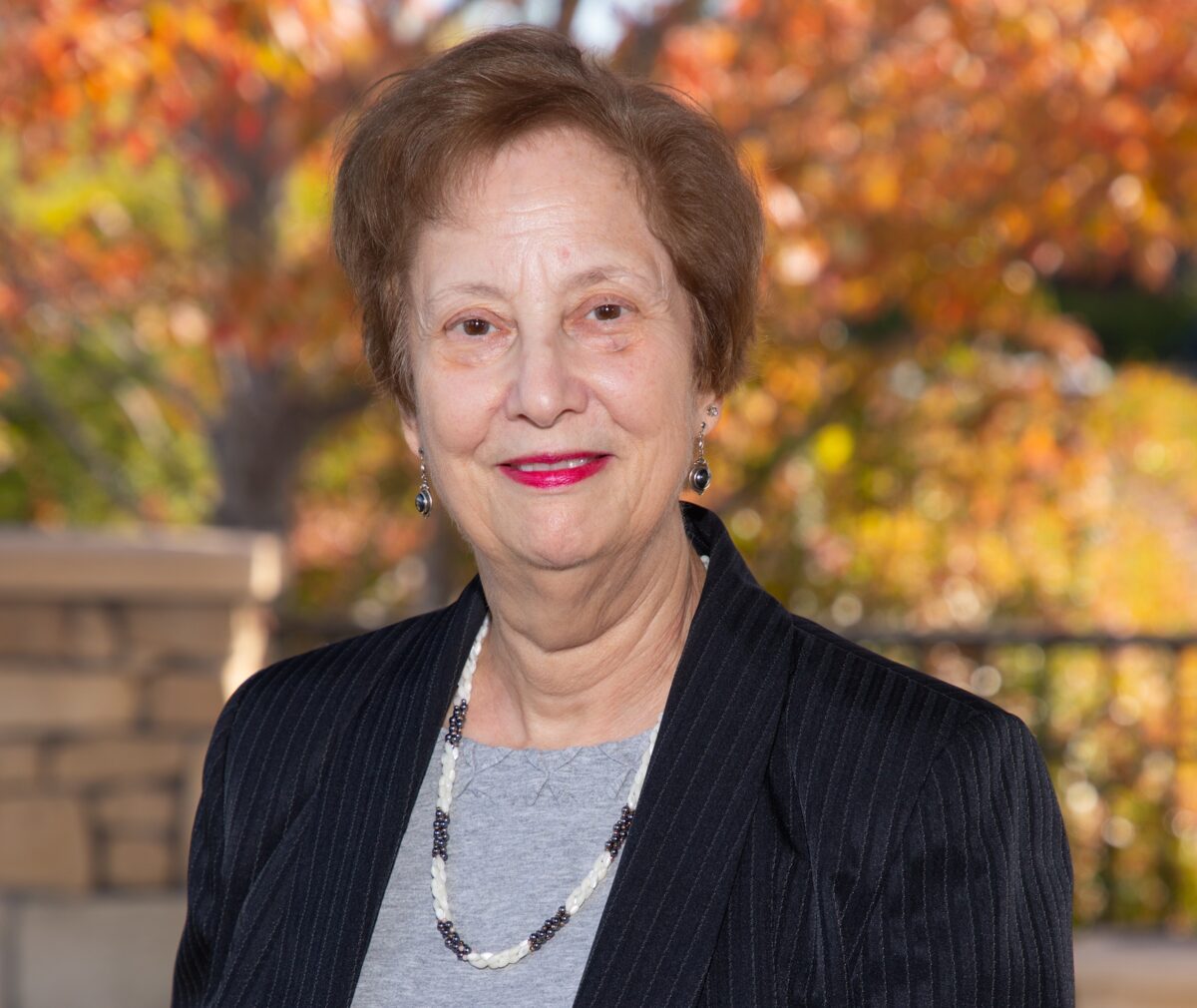 Portrait of an older woman with short auburn hair, smiling at the camera. She wears a gray top under a black pinstriped blazer, aong with a beaded black-and-white necklace and dangling earrings. She's standing outdoors with blurred atumn foiliage in orange, yellow, and green tones in the background.