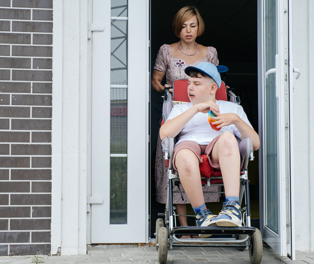 Mother with disabled teenage child in big stroller. Accessibility of people in wheelchairs. Woman going though the door, People with special Needs. Boy with Cerebral Palsy in chair going outside