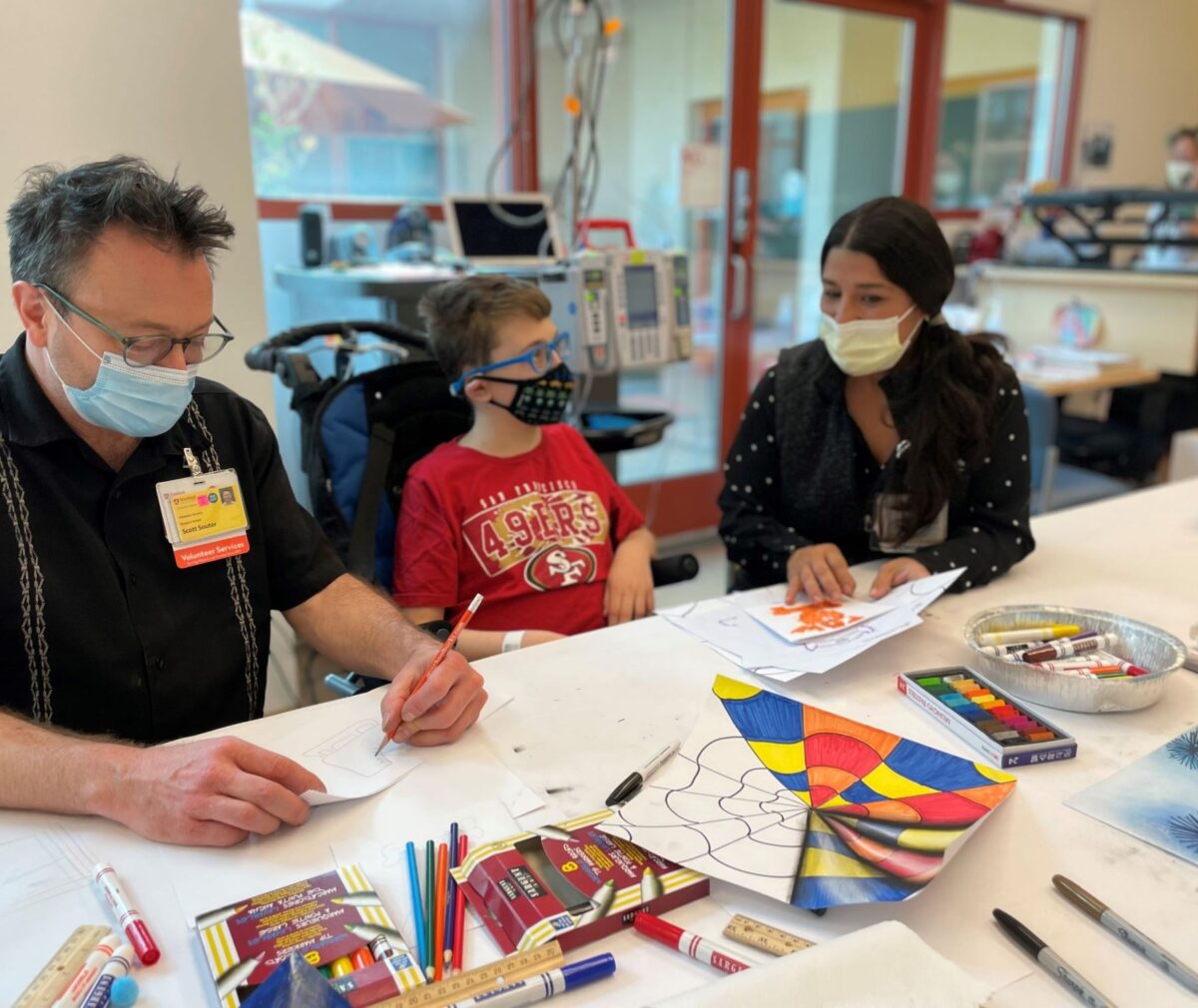 A masked hospital volunteer sketches on paper at a table across from a young boy in a 49ers T-shirt and a masked woman, both also masked, with colored pencils, markers, and a colorful geometric drawing spread between them; medical equipment and a wheelchair are visible in the background.
