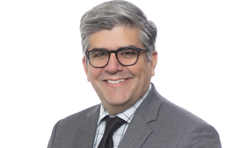 Headshot of a smiling man with gray hair and dark-framed glasses, wearing a gray suit, plaid shirt, and black tie, photographed against a white background.