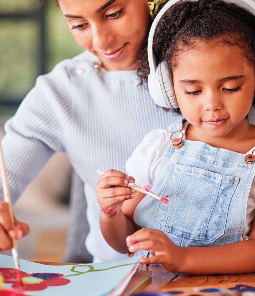 A woman helps a young girl wearing headphones paint a flower with red, blue, and yellow paint at a table.