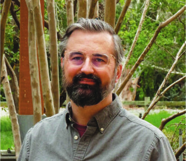 Headshot of a man with glasses, gray hair, and a dark beard wearing a gray button-down shirt, standing outdoors in front of trees and greenery.