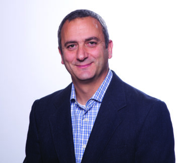 Headshot of a smiling man with short gray hair wearing a blue checkered shirt and dark blazer, against a light background.