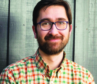 Headshot of a smiling man with dark hair, glasses, and a beard, wearing a colorful plaid shirt, standing against a wood plank background.