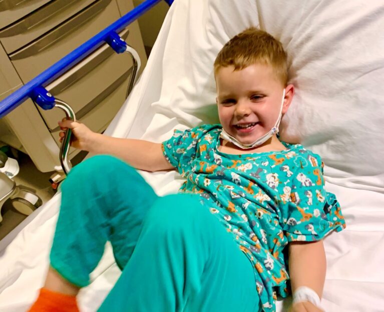 A young boy smiling in a hospital bed, wearing a teal patterned hospital gown and orange grip socks, with a nasal cannula and hospital ID bracelet on his wrist, playfully kicking up his leg.