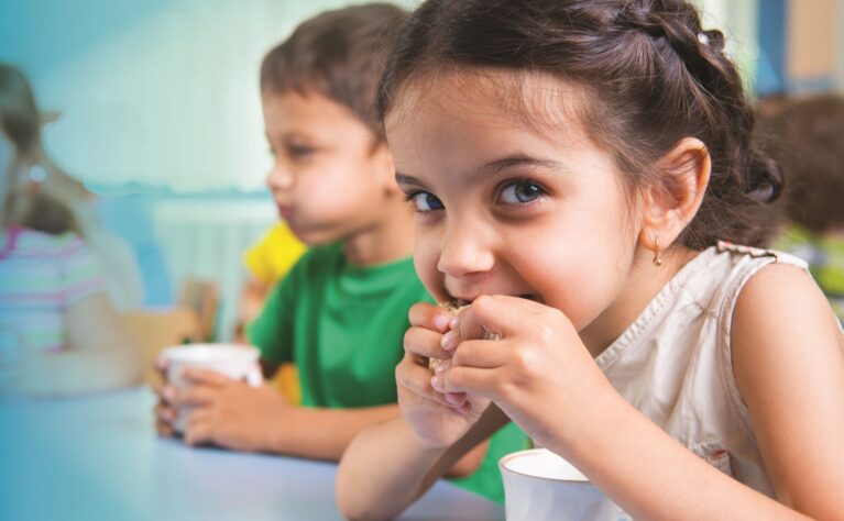 A young girl with braided dark hair eating a snack at a table, looking toward the camera, with other children seated nearby holding cups