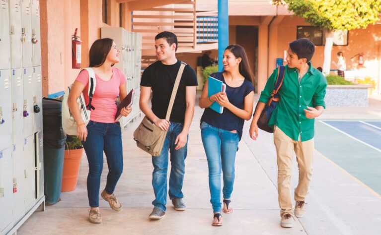 Four teenage students walking and talking together outdoors past a row of lockers on a school campus, carrying backpacks and notebooks