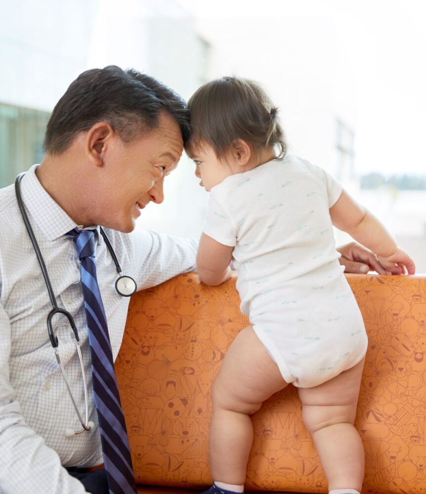 A doctor wearing a stethoscope smiles while leaning his forehead close to a baby standing on a patterned couch, facing away from the camera