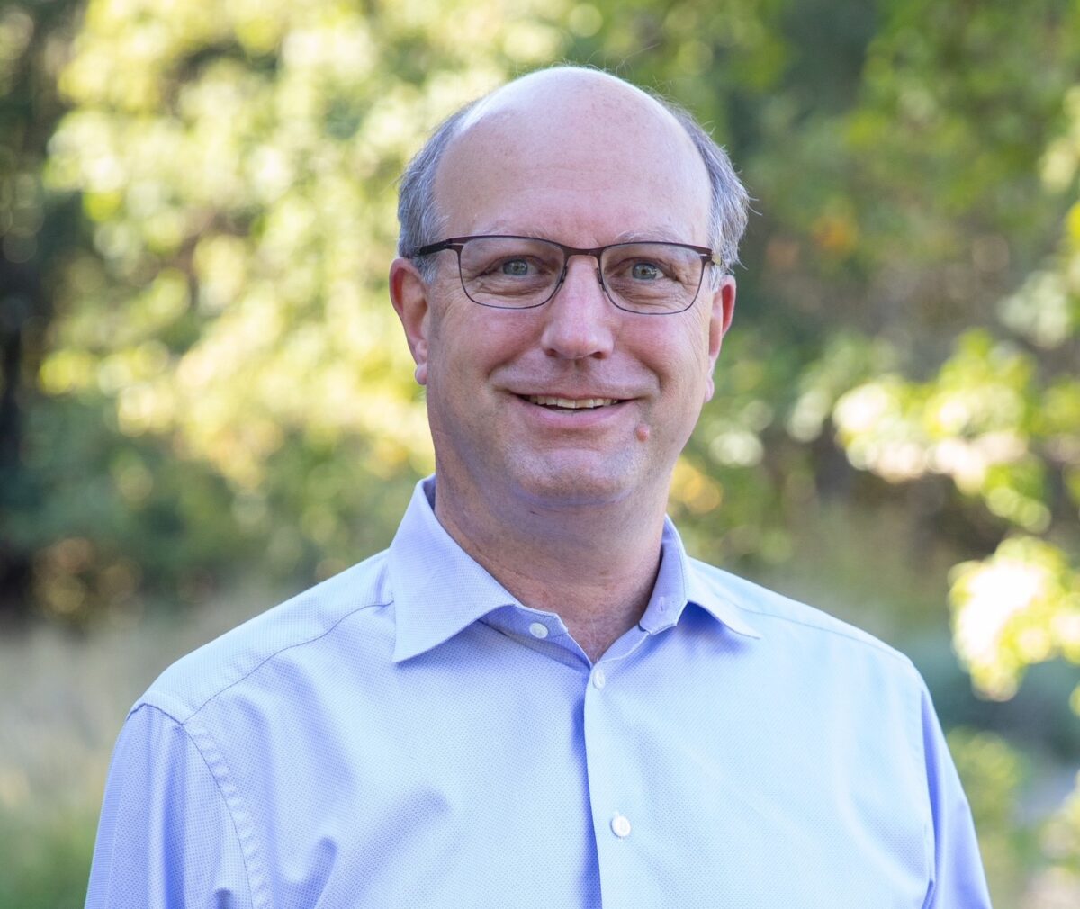 professional-style portrait photo of a man with glasses and a light blue button-down shirt, smiling outdoors with green trees blurred in the background.