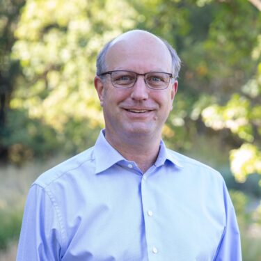 professional-style portrait photo of a man with glasses and a light blue button-down shirt, smiling outdoors with green trees blurred in the background.