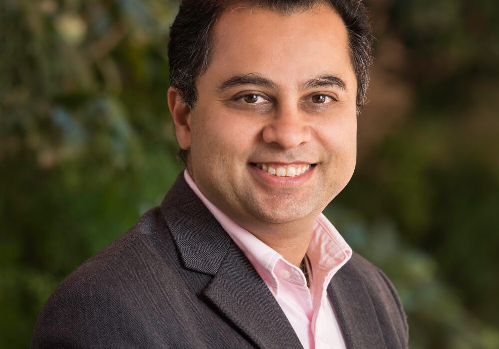 A man with dark hair, wearing a pink button-down shirt under a charcoal herringbone blazer, smiling with an outdoor green background similar to the first photo.