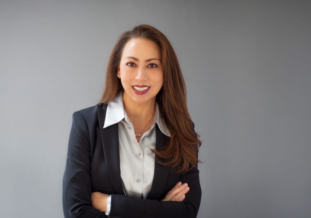 Professional studio headshot of a smiling woman with long brown hair, wearing a dark blazer and light-colored blouse, arms crossed, against a gray background.