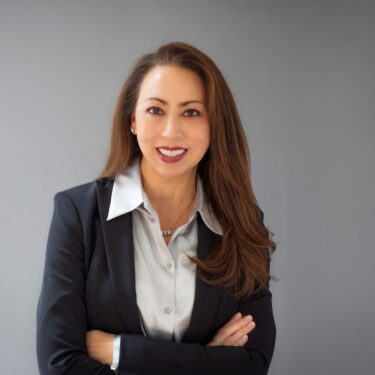 Professional studio headshot of a smiling woman with long brown hair, wearing a dark blazer and light-colored blouse, arms crossed, against a gray background.