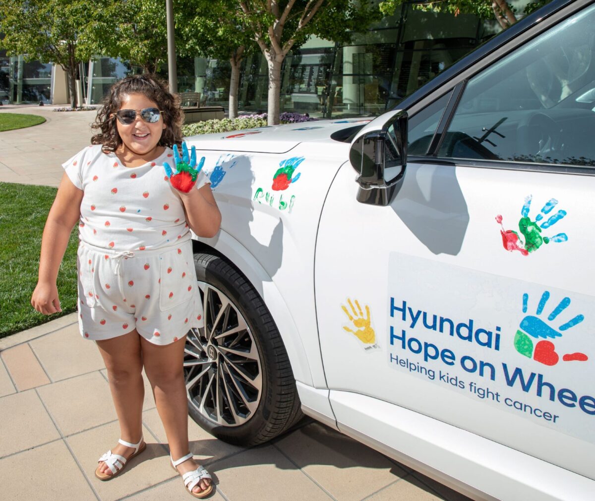 A young girl with curly hair and sunglasses stands next to a white car, holding up her hand freshly painted in blue, green, and red to make a handprint. The car door is decorated with colorful handprints and a 'Hyundai Hope on Wheels — Helping kids fight cancer' logo. She wears a white strawberry-print outfit and stands on a paved walkway with trees and a building in the background.