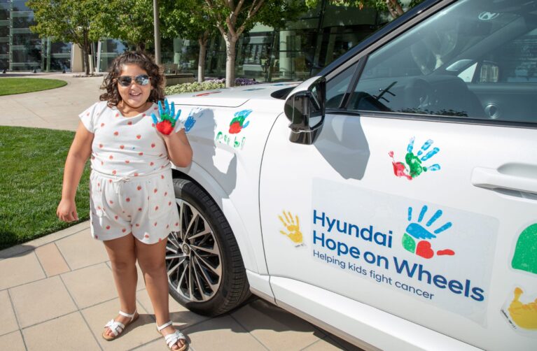 A young girl with curly hair and sunglasses stands next to a white car, holding up her hand freshly painted in blue, green, and red to make a handprint. The car door is decorated with colorful handprints and a 'Hyundai Hope on Wheels — Helping kids fight cancer' logo. She wears a white strawberry-print outfit and stands on a paved walkway with trees and a building in the background.