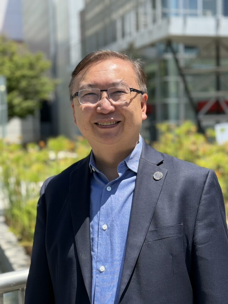 A smiling man with short dark hair and glasses, wearing a navy blazer over a light blue button-down shirt with a lapel pin, stands outdoors on a sunny day. Blurred greenery and a modern glass building are visible in the background