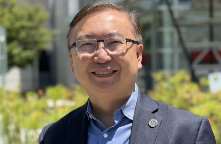 A smiling man with short dark hair and glasses, wearing a navy blazer over a light blue button-down shirt with a lapel pin, stands outdoors on a sunny day. Blurred greenery and a modern glass building are visible in the background