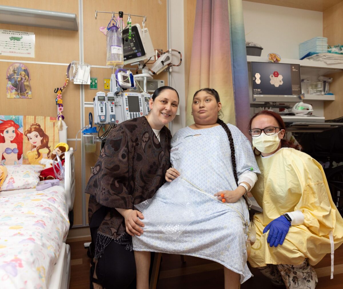 A young patient in a hospital gown sits between two smiling women in a hospital room decorated with Disney princess decor, including bedding, wall decals, and plush dolls. One woman wears a mask, gloves, and a yellow protective gown, while the other wears a patterned cardigan. Medical equipment, including IV poles and monitors, is visible in the background