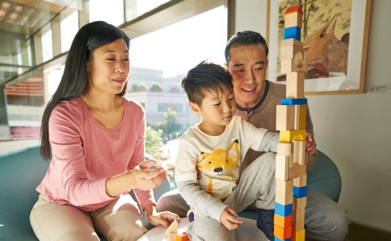 A child stacks colorful wooden blocks into a tall tower while two adults sit close by, smiling and watching. They're seated on a teal couch near a bright window with a hospital building visible outside.