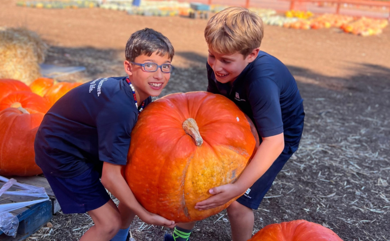 Two young children in blue shirts and shorts work together to lift a large, heavy pumpkin. One smiles at the camera while the other smiles down at the pumpkin.