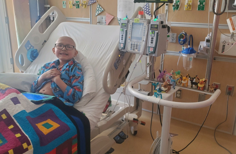 Lucile Packard Children's Hospital Stanford patient Josh wears a hospital gown and sits up right in a hospital bed. Josh smiles at the camera.