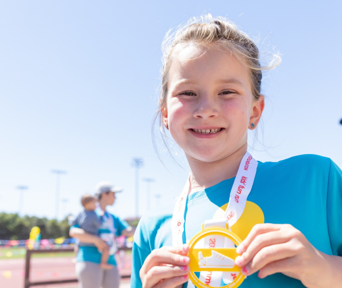 Smiling child in a teal Summer Scamper shirt holds up a yellow "kids' fun run" medal with a running shoe on it, on a sunny day at an outdoor track.