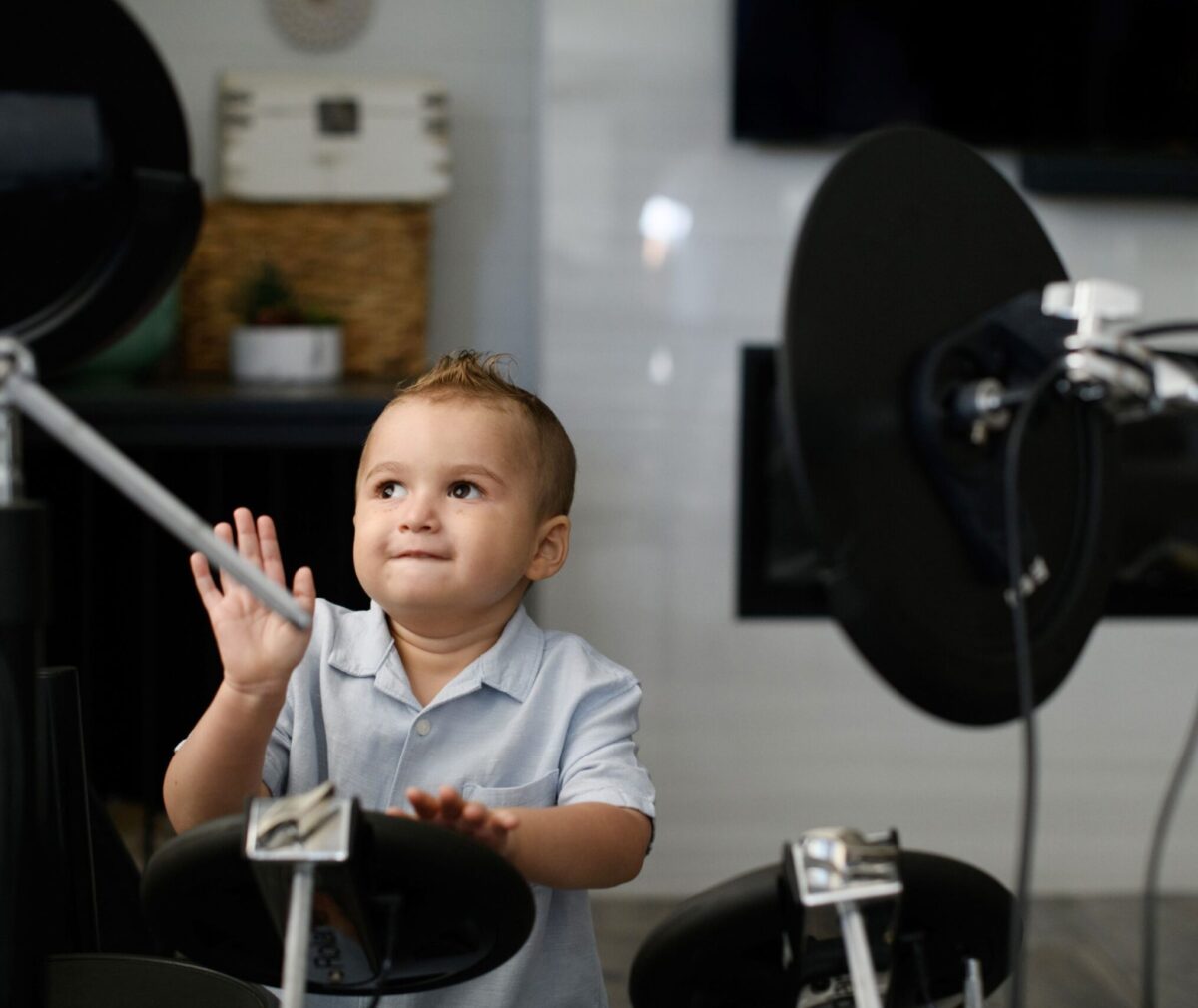 Toddler in a light blue button-up shirt sits behind an electronic drum kit, looking up with a curious expression as he raises one hand.