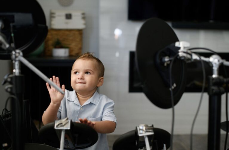 Toddler in a light blue button-up shirt sits behind an electronic drum kit, looking up with a curious expression as he raises one hand.