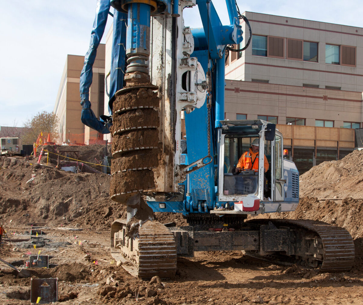 A large construction truck with a drill working in a dirt lot with the hospital in the background.