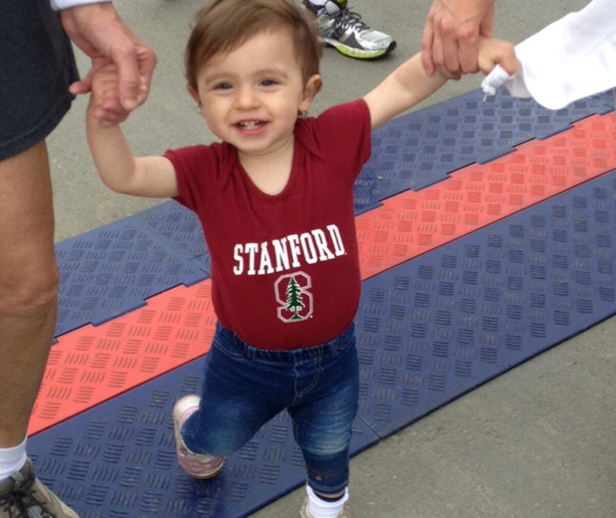 A toddler smiles in a Stanford University t-shirt as they hold the hands of two adults.