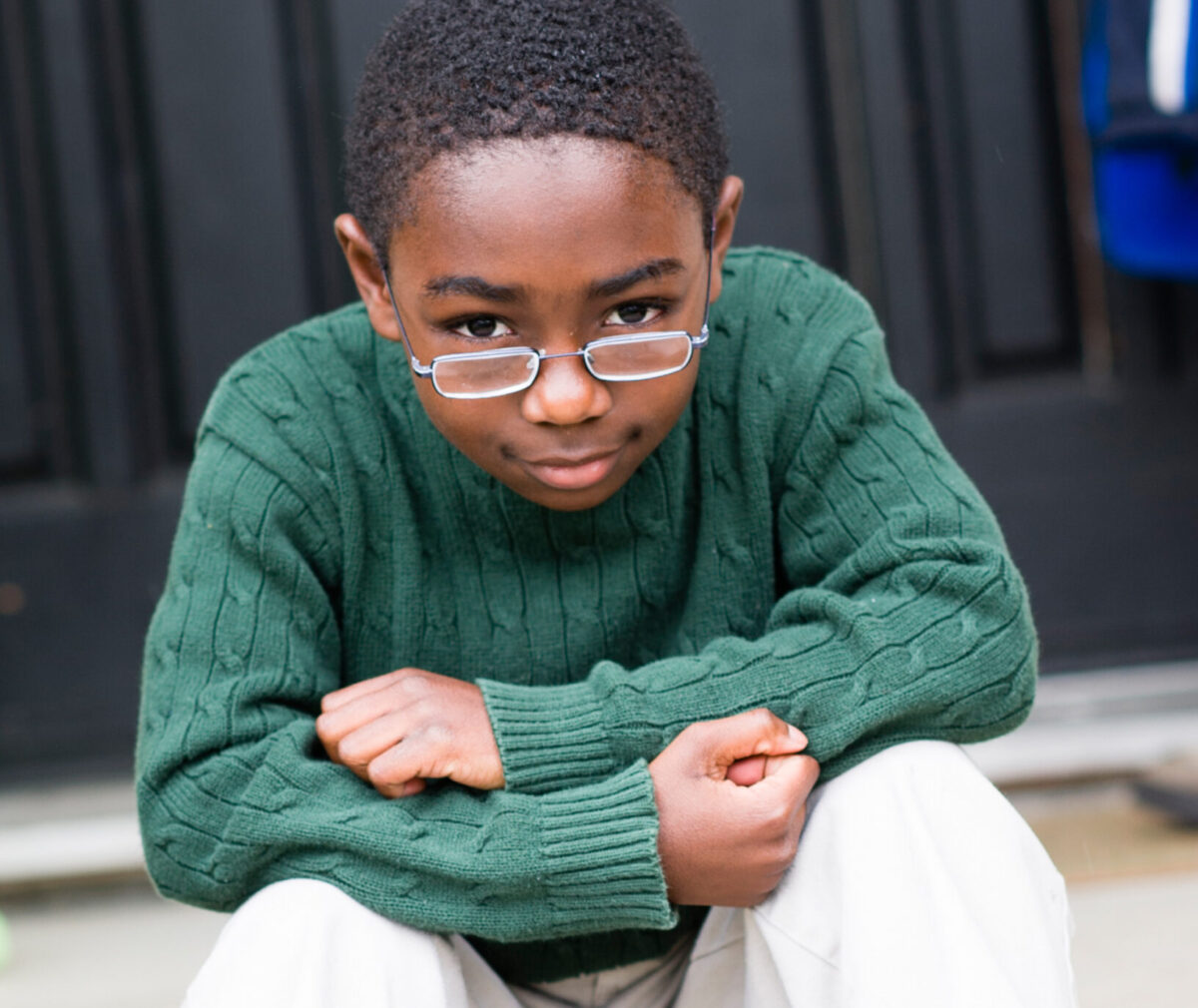 A young boy in a green sweater and glasses looks sad sitting on the front steps.