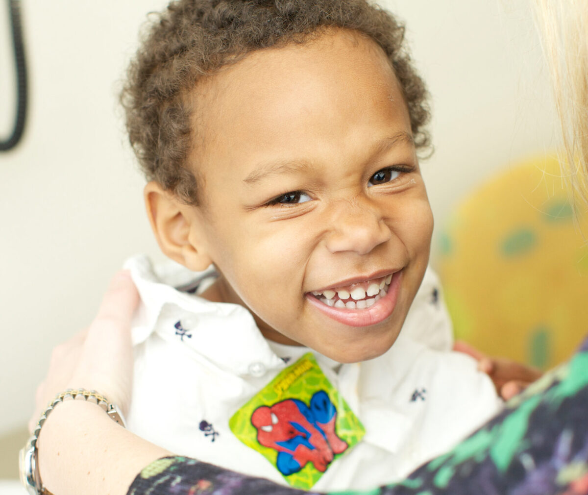 A young child smiles at the camera wearing a collared shirt with a spider man sticker on it.