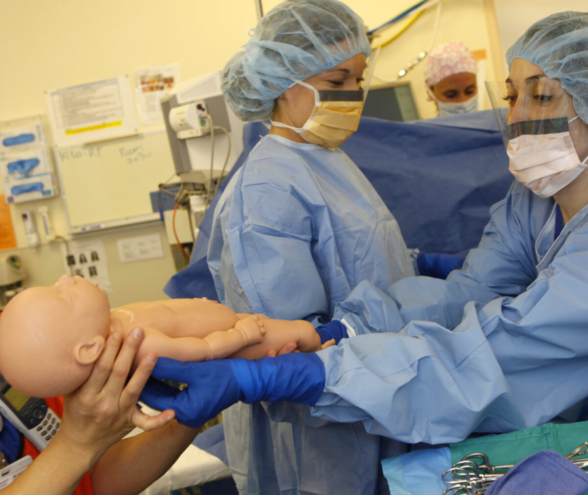 Three people in blue hospital scrubs, hair covers, and masks hand a mannequin baby across a busy hospital room.