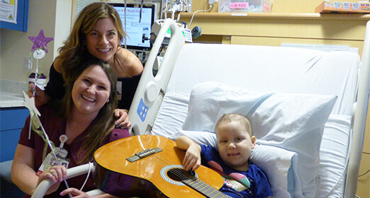 Child patient in hospital bed holding a guitar next to two adults.